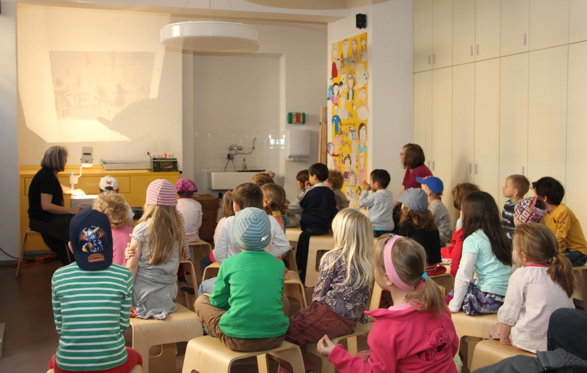 Lesung für Kinder Das Foto zeigt, wie eine Kindergruppe bei einer Lesung mit Bildern und Musik zuhört.