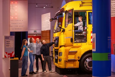 On the road again! Die Welt unserer Trucker Ein Junge sitzt am Steuer eines Lkw. Vier weitere Personen stehen vor dem Lkw und schauen zu ihm hinauf.