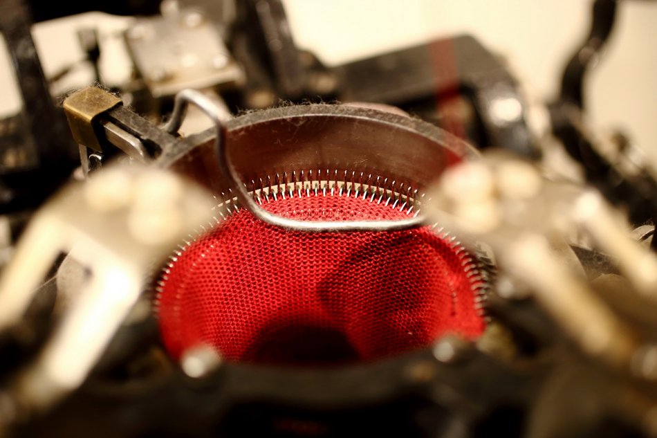 Das Foto zeigt eine Detailaufnahme einer historischen Kleinrundstrickmaschine. Die Nahaufnahme zeigt die komplexe Mechanik des Rundstrickwerks mit unzähligen, kreisförmig angeordneten Nadeln. Das im Beispiel verwendete Garn ist von roter Farbe.