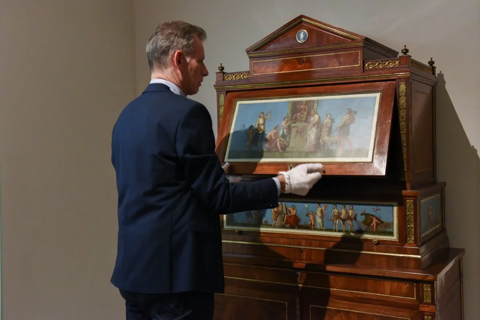 Ein Mann ist mit dem Rücken zu uns gekehrt und öffnet einen antiken Sekretär.