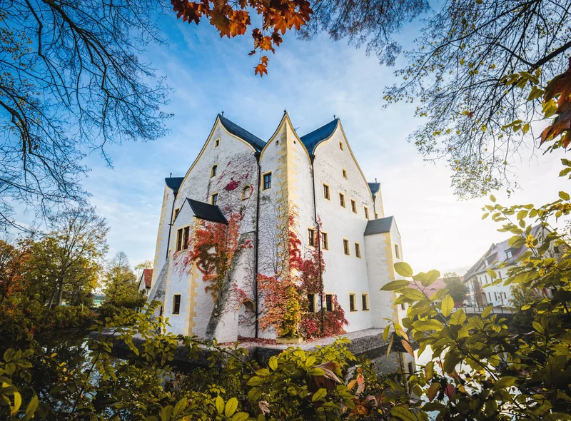 Blick über einen von Herbstlaub umgebenen Wassergraben auf ein kleines, weiß getünchtes Renaissance-Schloss