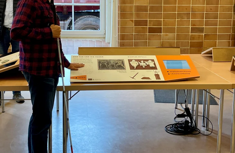 Ein sehbehinderter Mann mit Brille und Langstock steht vor einer Inklusivstation und liest mit einer Hand die auf ihr befindliche Braille-Schrift