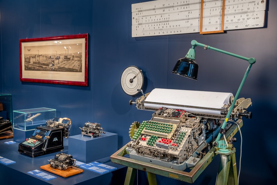 Eine historische Rechenmaschinen mit vielen grünen und einigen roten Tasten, kompliziert aussehender Mechanik und oben eingespannter Papierrolle steht neben weiteren Exponaten früher Rechentechnik vor einer blauen Ausstellungswand. An der Wand hängen ein überdimensionaler Rechenschieber und eine historische Grafik eines Industrie-Areals aus dem 19. Jahrhundert.