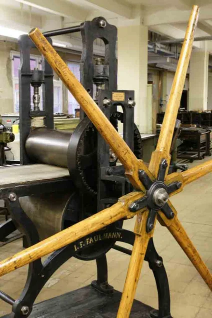In einem Saal steht eine historische Druckerpresse aus schwarzem Stahl mit großem hölzernen Antriebsrad.