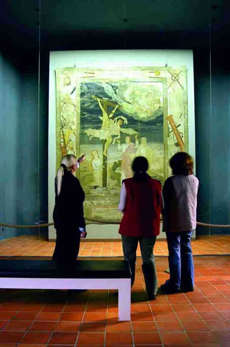 Drei Menschen sind von hinten zu sehen, wie sie auf eine große, rechteckige textile Arbeit blicken, die gut ausgeleuchtet vor einer dunklen Wand hängt. Die Motive auf dem Textil sind nicht im Einzelnen erkennbar.