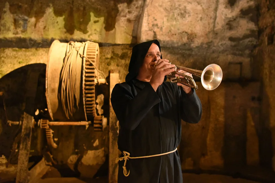Ein Mann in Mönchskleidung spielt Trompete. Er steht vor der Wagenwinde in der Dunklen Appareille der Festung Königstein.