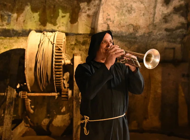 Ein Mann in Mönchskleidung spielt Trompete. Er steht vor der Wagenwinde in der Dunklen Appareille der Festung Königstein.