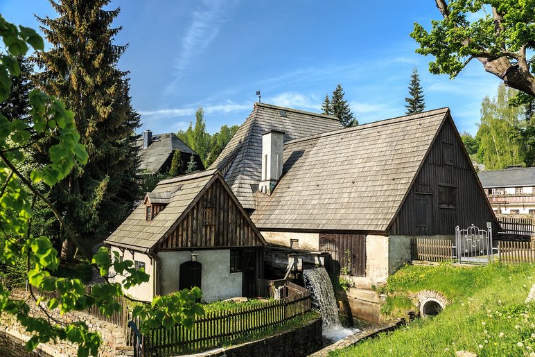 Ein Ensemble alter Häuser mit hölzernen Spitzdächern stehen in grüner Landschaft, zwischen zwei der Häuser verläuft ein Bach, der von einem Wasserrad unterbrochen wird.