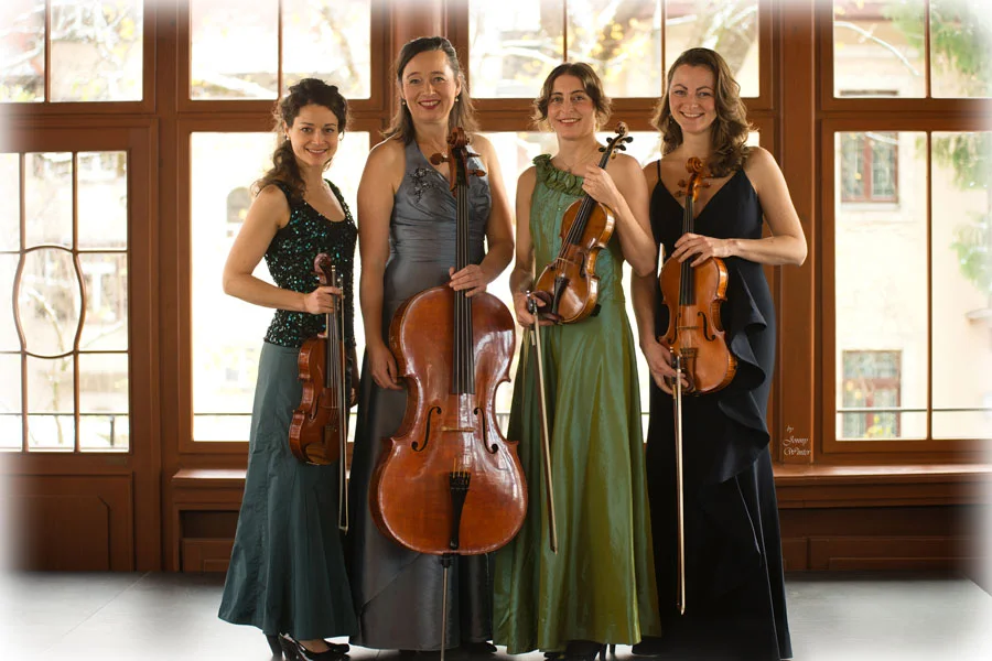 Lunus-Quartett, Dresden Vier nebeneinanderstehende junge Musikerinnen mit ihren Streichinstrumenten stehen in einem Raum vor einer großen Fensterwand und lächeln in die Kamera.