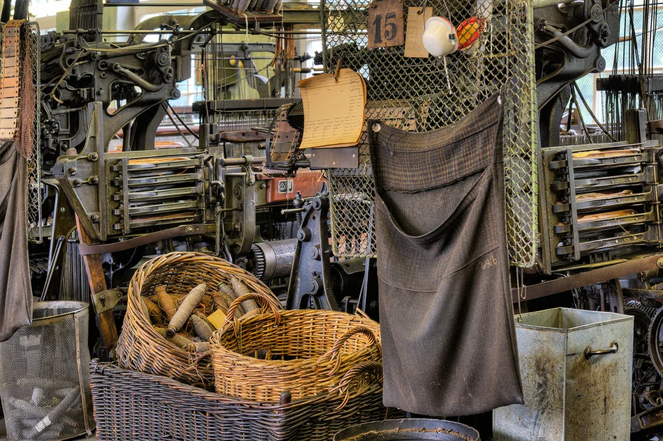 Weberei: Historische Webstühle Mehrere alte Webmaschinen stehen hinter- beziehungsweise nebeneinander, davor stehen Bastkörbe mit Garnspindeln.