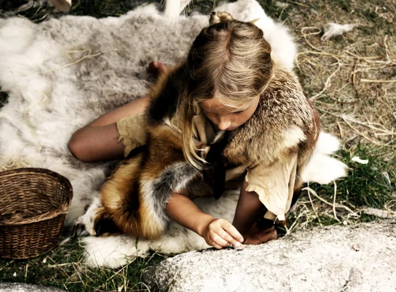 Ein Kind in Fellkleidung sitzt neben einem geflochtenen Korb auf einem Tierfell.