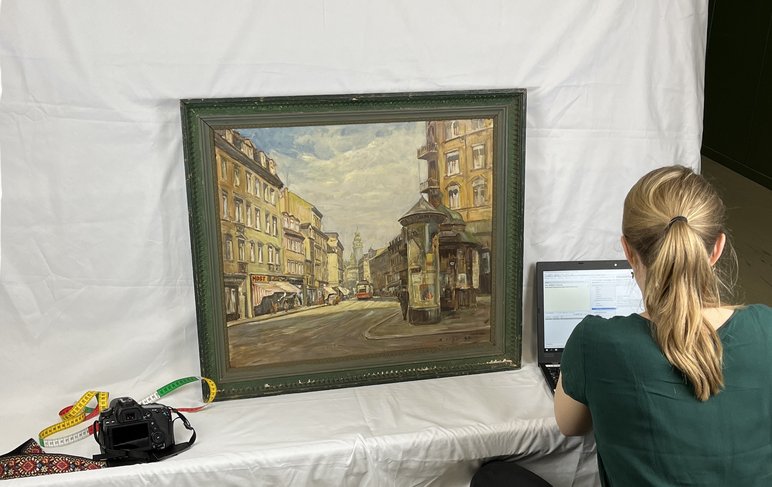 Auf einem mit weißem Stoff bedeckten Tisch steht das Gemälde einer Leipziger Stadtansicht, links daneben liegen eine Kamera und ein Maßband. Rechterhand sitzt eine Frau an einem Laptop.