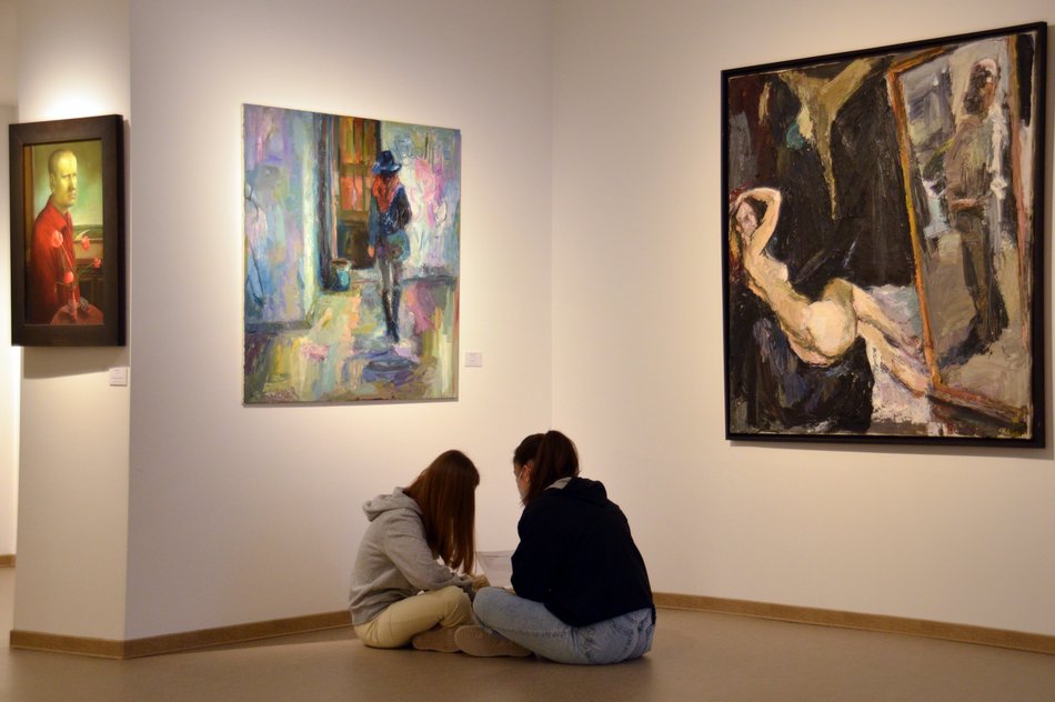 Auf dem Foto sind zwei jugendliche Mädchen von hinten zu sehen, die, auf dem Boden sitzend, sich über die an der Wand hängenden Gemälde mit Darstellungen menschlicher Körper beziehungsweise Gesichter austauschen. In den Händen halten sie Clipboards.
