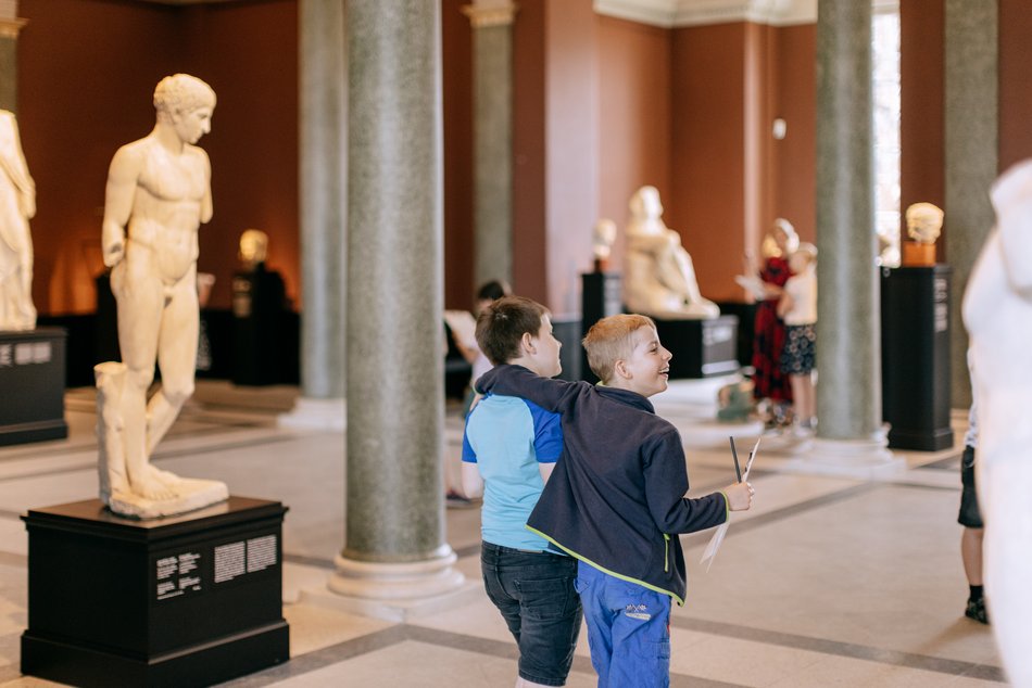 Das Foto zeigt zwei Schüler bei einem Vermittlungsprogramm in der Skulpturensammlung bis 1800 der Staatlichen Kunstsammlungen Dresden. Sie laufen durch eine große Säulenhalle mit mehreren antiken Skulpturen.