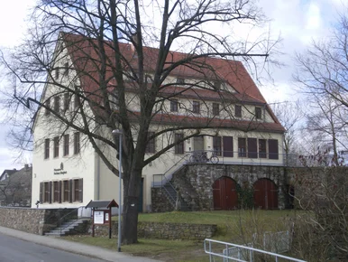 „Huthaus Einigkeit“, Brand-Erbisdorf Außenansicht eines dreistöckigen Gebäudes mit Spitzdach, das von einer Natursteinmauer umzäunt ist. Davor steht ein großer, jahreszeitlich bedingt kahler Baum.