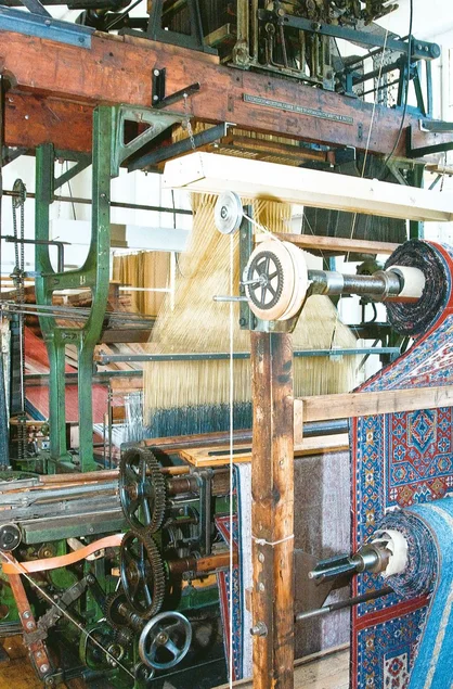 In einer großen historischen Maschine laufen unzählige Fäden zusammen und werden zu einem Teppich mit klassischem Muster verwebt