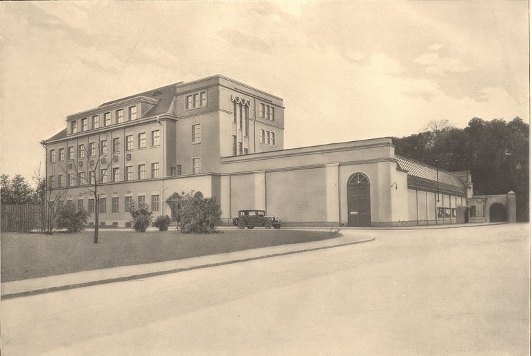 Sepia-farbenes historisches Foto eines an einem großen Platz stehenden viergeschossigen Gebäudes mit Flachdach, davor ein historisches Automobil