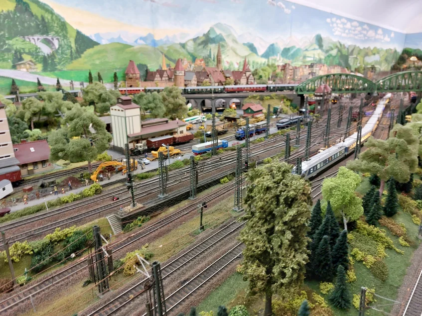 Foto eines mehrspurigen Bahnhofbereiches einer Modellbahnanlage mit Zügen, Gebäuden und einer gemalten Bergkulisse im Hintergrund
