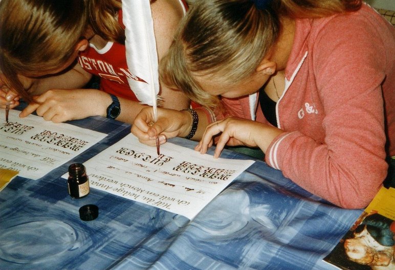 Zwei junge, eng nebeneinandersitzende Personen schreiben mit Federkielen in Frakturschrift auf Papier, vor ihnen steht ein kleines Tintenfass.