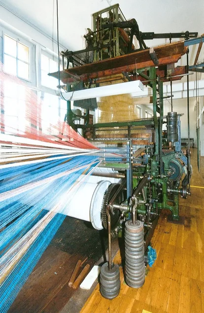 Unzählige blaue, weiße und rote Fäden laufen parallel auf eine große historische Webmaschine zu