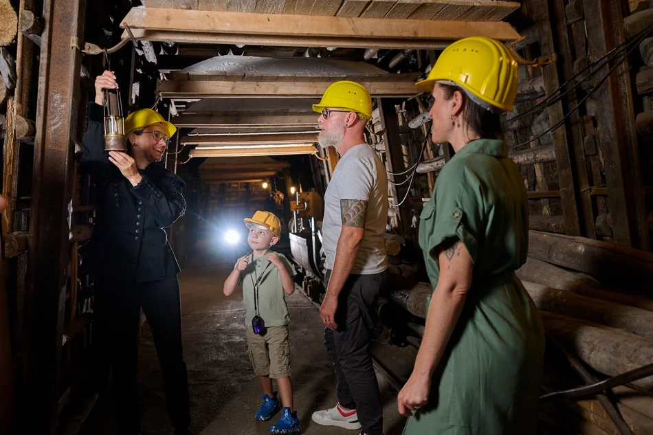 Anschauungsbergwerk Zwei Erwachsenen und einem Kind im Untertagebereich wird von einem Guide die Funktion einer Grubenlampe erklärt.