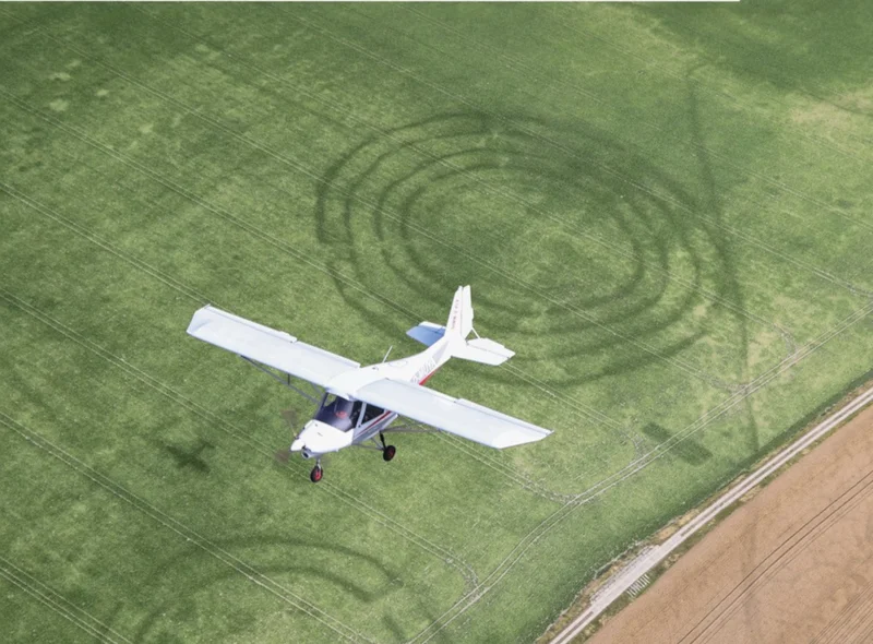 Luftbild, das ein über ein Feld mit kreisrunden Strukturen fliegendes Kleinflugzeug zeigt