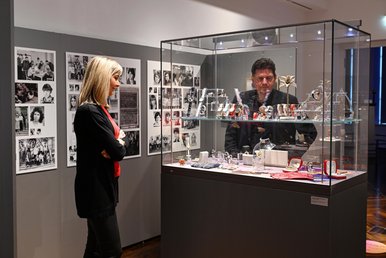 Blick in die Sonderausstellung „Die Zeit ist weiblich.“ Eine Frau und ein Mann blicken in eine mit einer Vielzahl von Kleinobjekten bestückte Vitrine. An der Wand dahinter hängen zahlreiche Schwarzweißfotos.