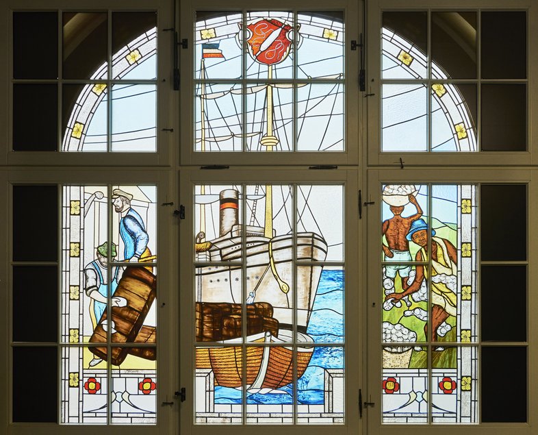Zentrales Motiv des historischen Bleiglasfensters ist ein Schiff, das von zwei europäischen Männern beladen wird. Am Ufer daneben sind zwei dunkelhäutige Männer beim Baumwollpflücken zu sehen.