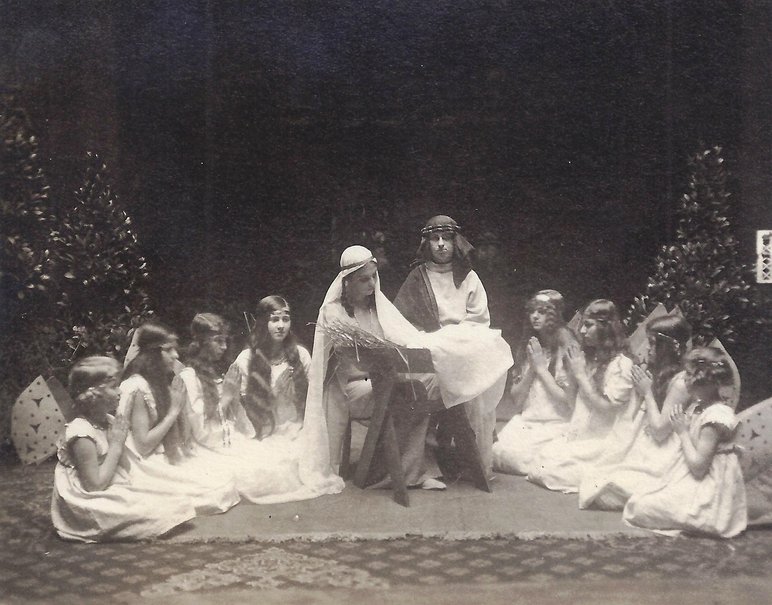 Schwarzweißfoto einer Krippenspiel-Aufführung: Maria und Josef blicken liebevoll in die Krippe, im Halbkreis um sie herum sitzen acht weißgekleidete Engel.