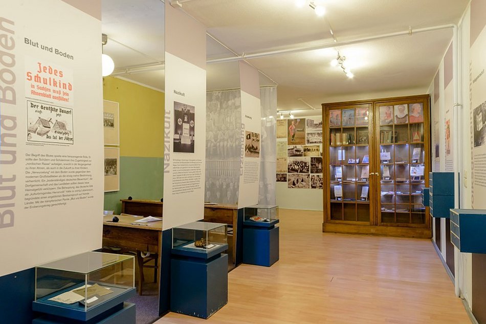 Foto eines Blicks in den Ausstellungsbereich zur Schule im Dritten Reich, mit einer Vielzahl von Text- und Bildbannern, einem als Vitrine dienenden großen Schrank mit Glastüren und historischen Schulbänken