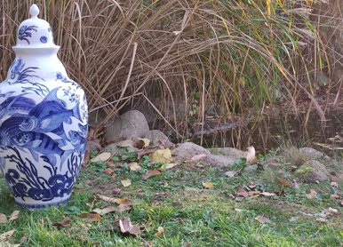 Eine weiße Vase mit Fisch- und Unterwassermotiven in Blautönen steht an der Uferböschung eines Gewässers