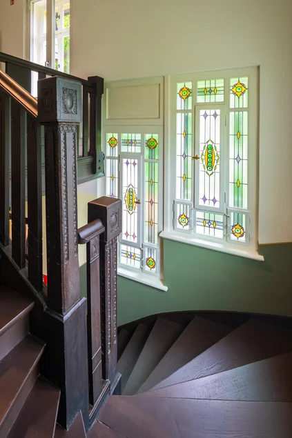 Eine Treppe aus dunklem Holz windet sich von einer Etage in die nächste, in die ebenfalls hölzernen Geländer sind Verzierungen geschnitzt. Die Treppenhausfenster bestehen aus kunstvollem buntem Glas im Stil der Gründerzeit.