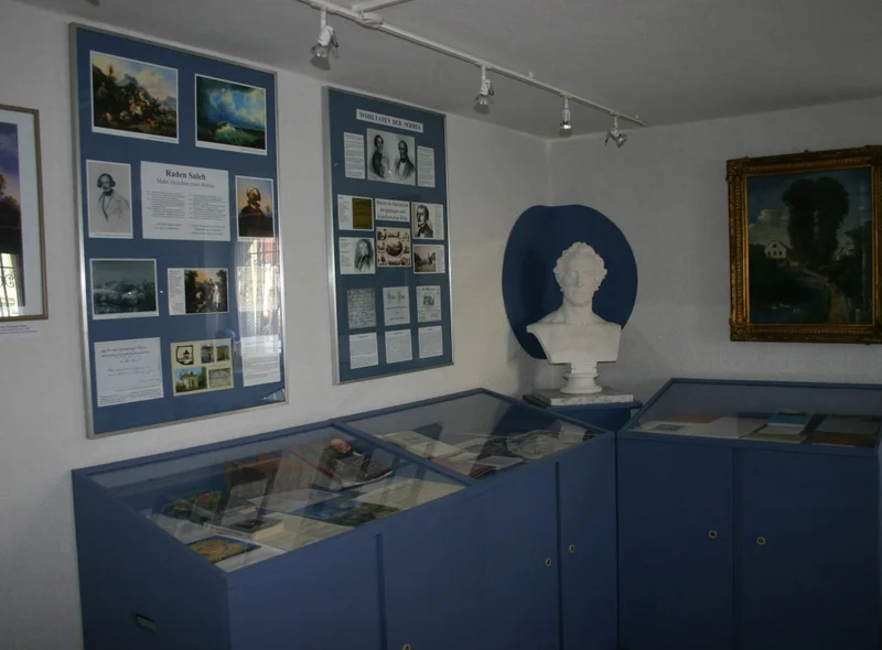 Blick in einen Ausstellungsraum mit Tischvitrinen, an dessen Wänden zwei Gemälde sowie zwei Infotafeln hängen. In der Ecke steht die weiße Büste eines Mannes.