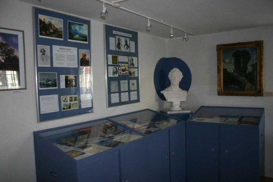 Blick in einen Ausstellungsraum mit Tischvitrinen, an dessen Wänden zwei Gemälde sowie zwei Infotafeln hängen. In der Ecke steht die weiße Büste eines Mannes.