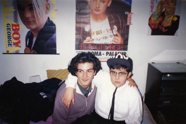 Farbfotografie zweier Jugendlicher, die Arm in Arm nebeneinander sitzen. Rechts von ihnen steht eine Stereoanlage, an der Wand hinter ihnen hängen drei Fotos des queeren Künstlers Boy George.