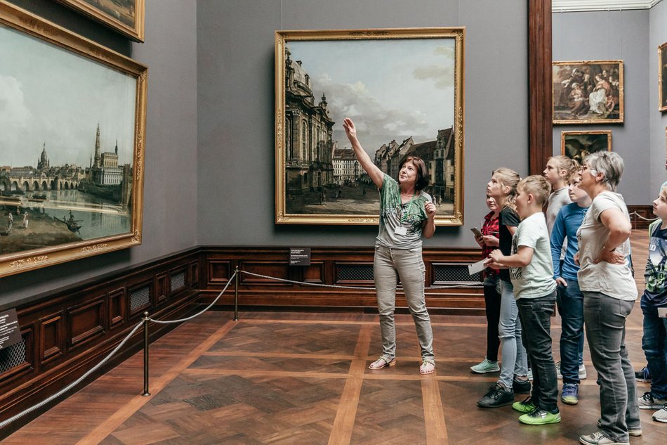Das Foto zeigt eine Gruppe von Schülerinnen und Schülern mit ihrer Lehrerin in einem Ausstellungssaal der Gemäldegalerie Alte Meister. Die vor ihnen stehende Kunstvermittlerin erläutert die Vedute „Dresden vom rechten Elbufer unterhalb der Augustusbrücke“, geschaffen 1748 vom venezianische Maler Bernardo Bellotto.