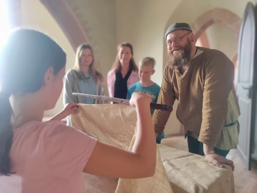 Das Maß aller Dinge – richtig messen früher und heute Ein in ein historisches Kostüm gekleideter Mann, zwei junge Frauen und ein Junge schauen einem Mädchen dabei zu, wie sie mit einer alten Holz-Elle ein Stück Stoff abmisst.