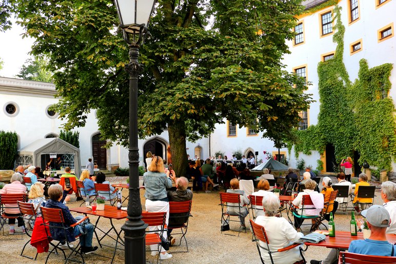 Viele Menschen sitzen in einem mit Café-Bestuhlung ausgestatteten Schlosshof. Unter einem großen Laubbaum geben Musiker ein Konzert.