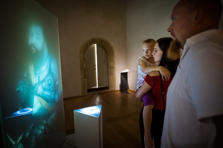 In einem dunklen Raum blicken ein Mann, eine Frau und ein Kind auf die digitale Animation eines mittelalterlich gekleideten Mannes.