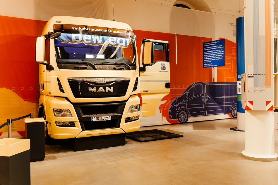 Die Fahrerkabine eines MAN-Lkw scheint eine Ausstellungswand zu durchbrechen.
