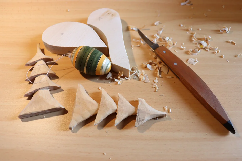 Schnitzen Auf einem Tisch liegen verschiedene Objekte aus Holz: zwei geschnitzte Bäumchen, ein gedrechseltes und bunt bemaltes Osterei, zwei Holzstücke die aneinandergelegt ein Herz ergeben. Daneben liegt ein Schnitzmesser. Um alles herum sind Holzspäne verteilt.