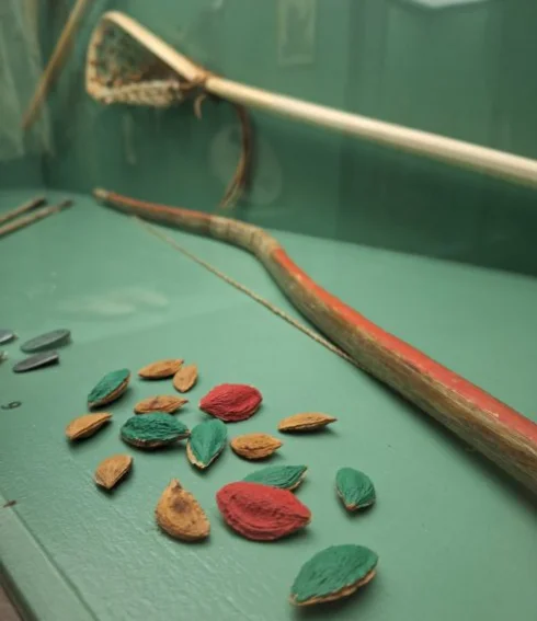 In einer Vitrine im Karl-May-Museum Radebeul liegen rot, grün und blau bemalte Obstkerne sowie bemalte Stöcke und ein Kescher