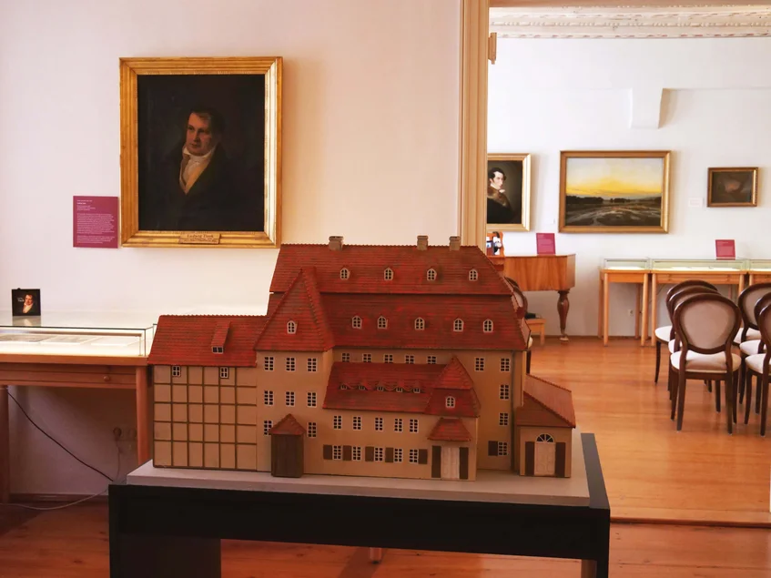 Zwei historische Ausstellungsräume mit dem Modell eines großen Gebäudes im Vordergrund und Gemälden im Hintergrund