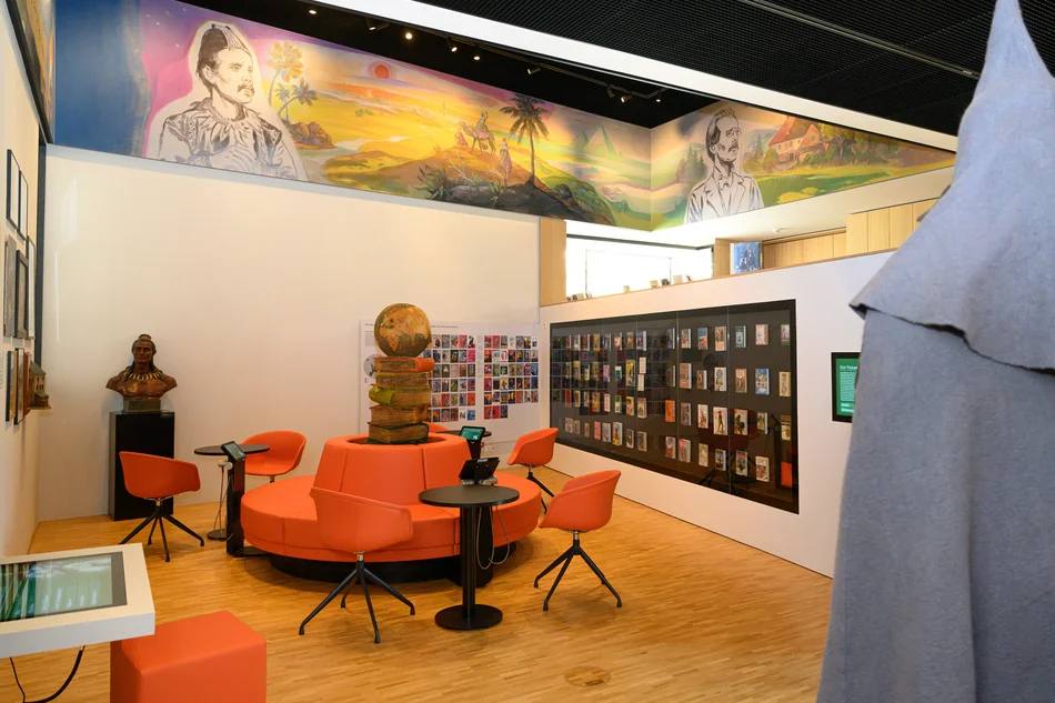 Barrierefreier Informationsplatz und Fremdsprachenbereich Das Foto zeigt einen barrierefrei gestalteten Informationsplatz, der aus einer kreisförmig angeordneten Sitzgruppe mit drei Medienstationen und drei Beistelltischen besteht. An zwei dahinter befindlichen Wänden wird eine Vielzahl von Buch-Covern präsentiert. Eine weitere, unterfahrbare Medienstation ist im Vordergrund links zu sehen.