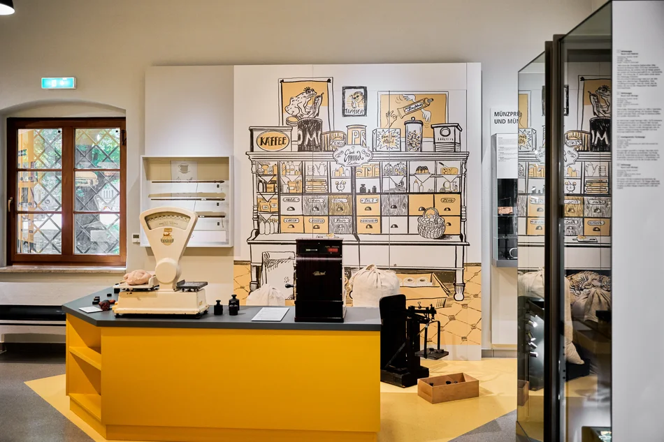 Foto der Inszenierung eines Krämerladens: Zentral auf der Theke befinden sich eine weiße Lebensmittelwaage mit mehreren schwarzen Gewichten sowie eine historische Kasse. Auf dem Boden hinter der Theke stehen eine historische schwarze Waage sowie prall gefüllte weiße Säcke. Die Rückwand ziert die grafische Darstellung eines alten Krämerladens mit Regalen und Schubkästen.