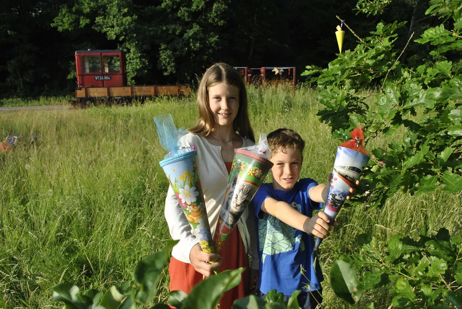 Ein Mädchen und ein Junge stehen auf einer grünen Wiese und halten Zuckertüten in die Kamera. Im Hintergrund ist ein Eisenbahnwaggon zu sehen.