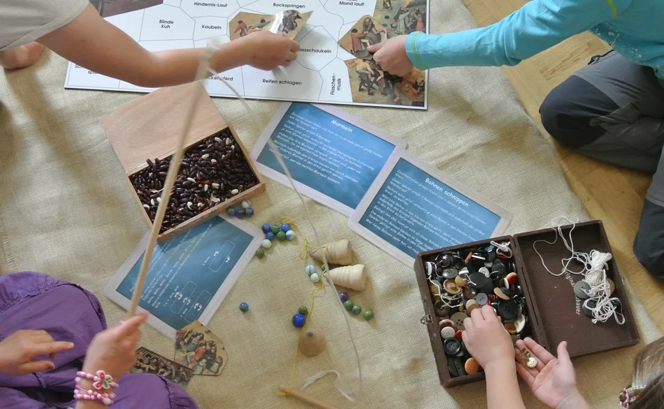 Draufsicht auf einen Sitzkreis, in dessen Mitte ein Brettspiel, Kreisel, ein Kistchen mit Knöpfen und verschiedene Fäden und Schnüre liegen