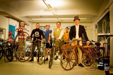 Im Foto sind sechs Männer in einem Raum zu sehen, die historische Fahrräder aus unterschiedlichen Zeiten präsentieren. Die Männer sind entsprechend der Fahrradmode der jeweiligen Zeit gekleidet.