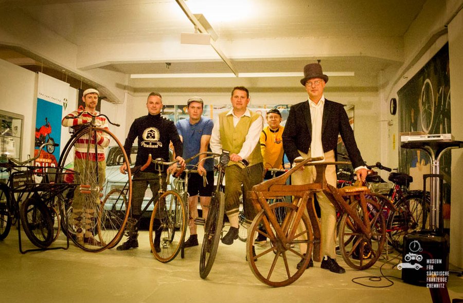 Im Foto sind sechs Männer in einem Raum zu sehen, die historische Fahrräder aus unterschiedlichen Zeiten präsentieren. Die Männer sind entsprechend der Fahrradmode der jeweiligen Zeit gekleidet.