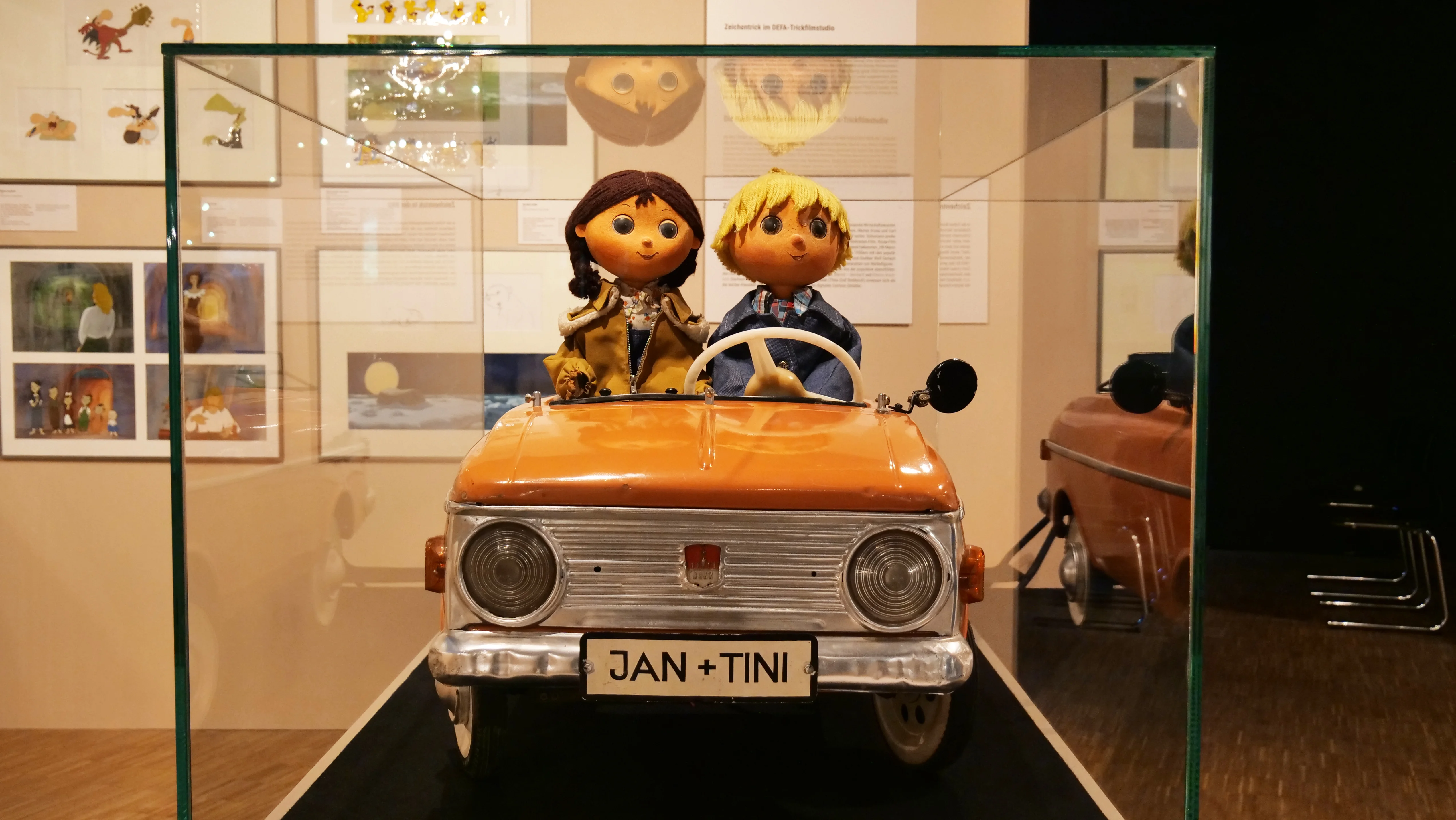 Zwei Puppen sitzen nebeneinander in einem Miniatur-Auto, auf dessen Nummernschild "Jan und Tini" steht.